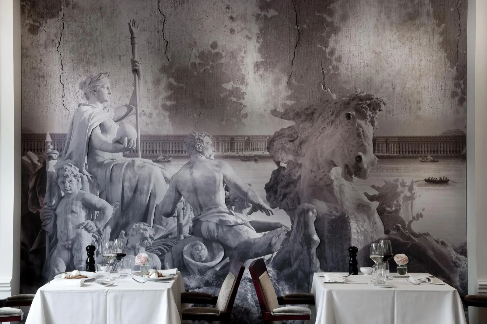 Elegant restaurant dining room with two dressed tables in front of a gray mural depicting mythological figures and horses, inspired by classical Bordeaux architecture.