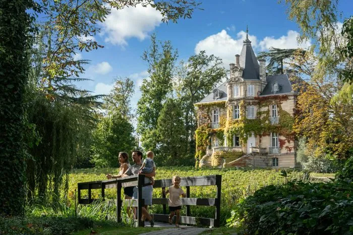 Family viewing a wine estate château from a wooden terrace overlooking the Graves vineyards near Bordeaux, with a Belle Époque mansion surrounded by lush greenery.