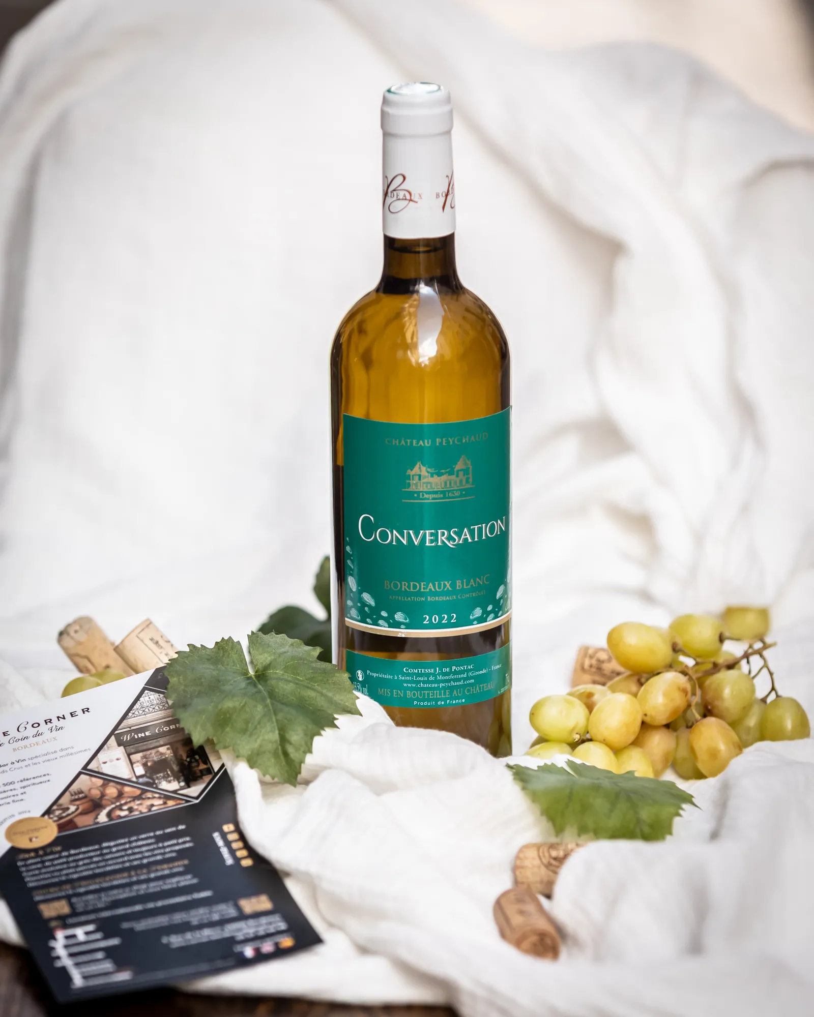 Conversation white wine bottle from Bordeaux 2022 surrounded by green grapes, vine leaves and cork stoppers on white fabric with Wine Corner documents in the background