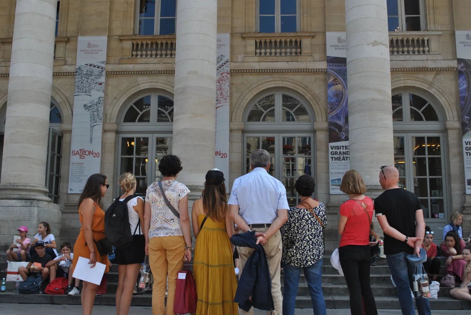 A group of visitors waits in front of the Bordeaux National Opera, a classical building with white columns and arched windows, with cultural programming banners displayed.