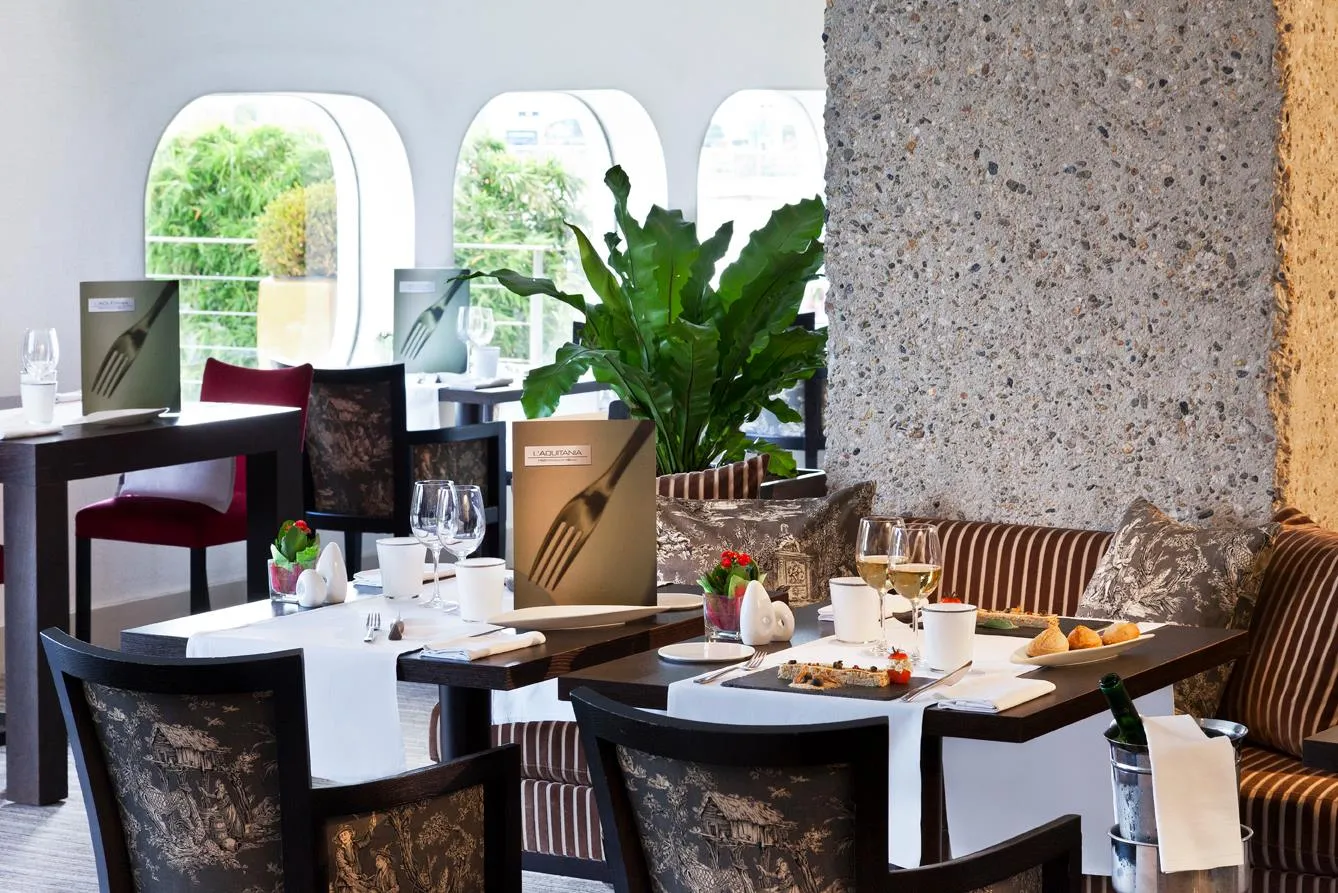 Elegant dining room with set tables, black and gold velvet chairs, arched windows overlooking greenery, floral and minimalist decor.
