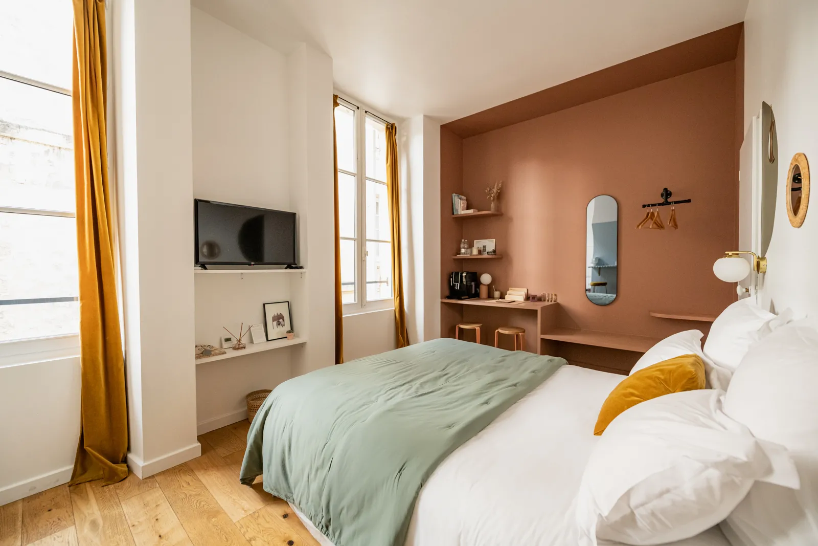 Modern bedroom with double bed, terracotta accent wall, wall-mounted TV, oval mirror, built-in desk and windows with golden yellow curtains at La Maison Fernand in Bordeaux