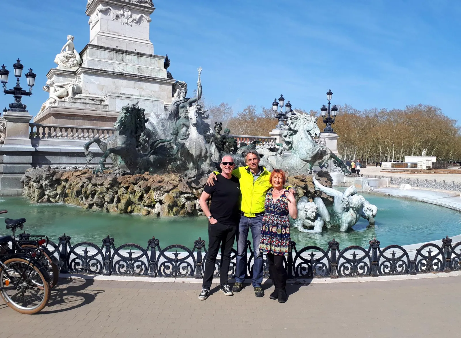 Three people posing in front of the Fountain of the Girondins in Bordeaux, featuring equestrian statues and a monumental column in the background, during a cycling tour.