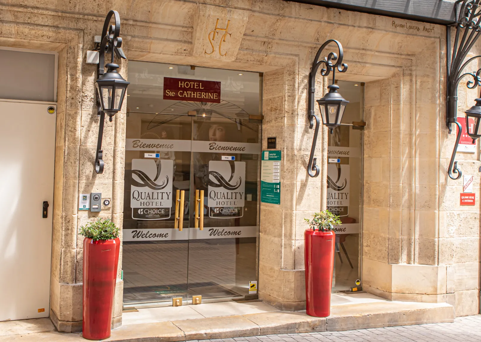 Entrance facade of Hôtel Sainte Catherine, a Quality Hotel in Bordeaux, featuring glass doors, wrought iron wall lanterns, and red welcome planters