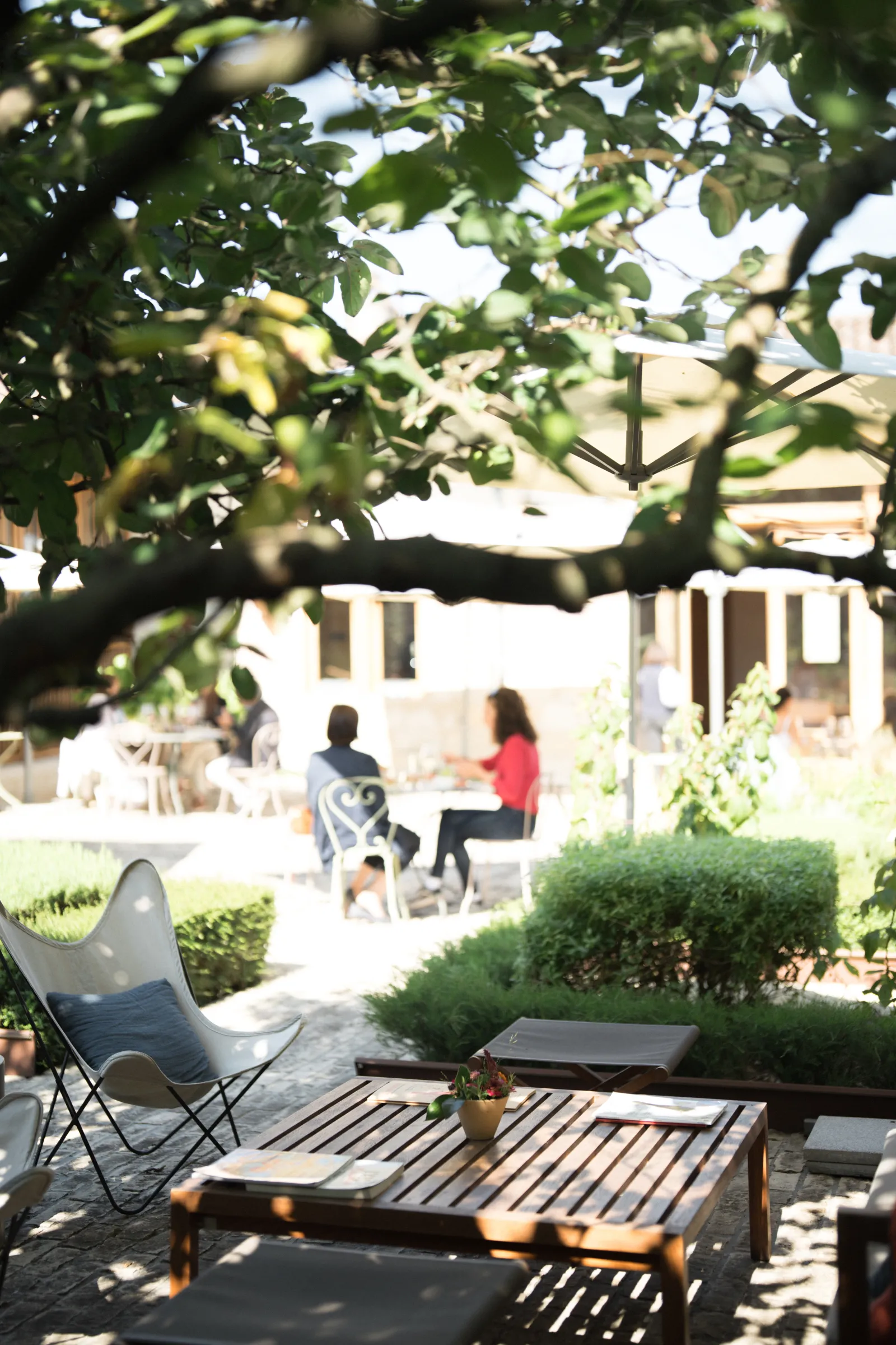 Shaded terrace at Château Marquis d'Alesme in Bordeaux featuring a lounge chair in the foreground and two people seated at a table under the trees in the background