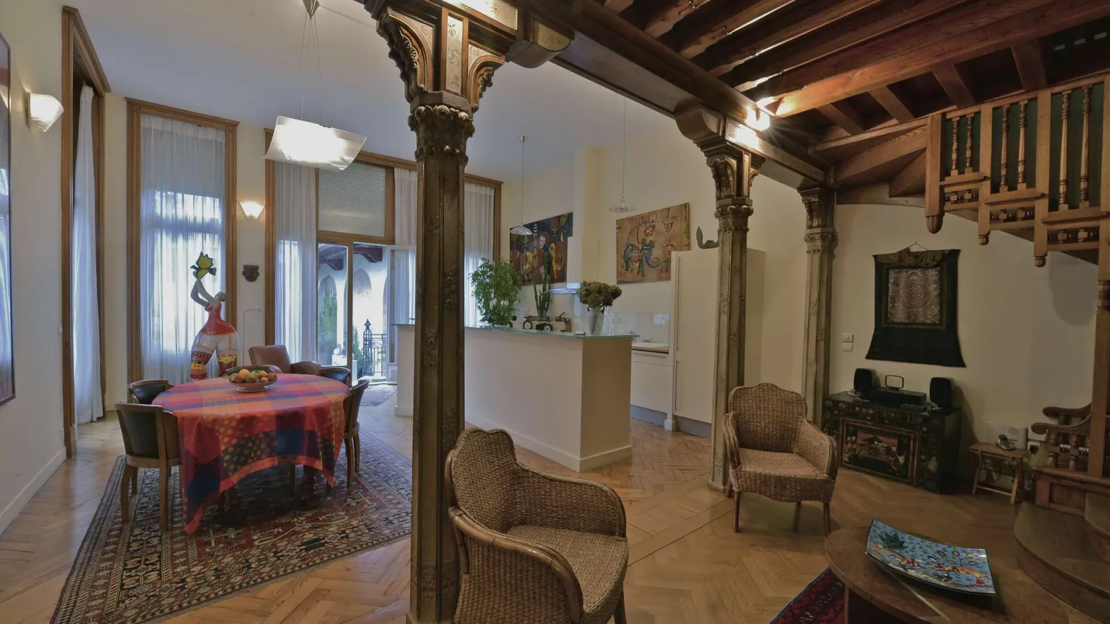 Interior of the Hôtel Frugès in Bordeaux: spacious living area with ornamental columns, rattan furniture, round table with colorful patterns, integrated kitchen, exposed wooden ceiling beams, and large windows opening onto a terrace.