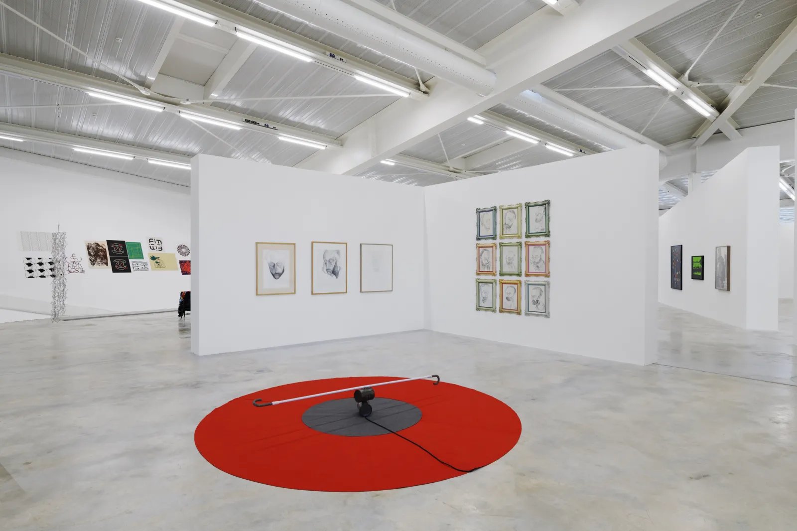 Contemporary art gallery interior featuring white partition walls, framed artworks on display, a large red circular disc on the floor with a central installation piece, and industrial ceiling with metal beams and linear lighting.