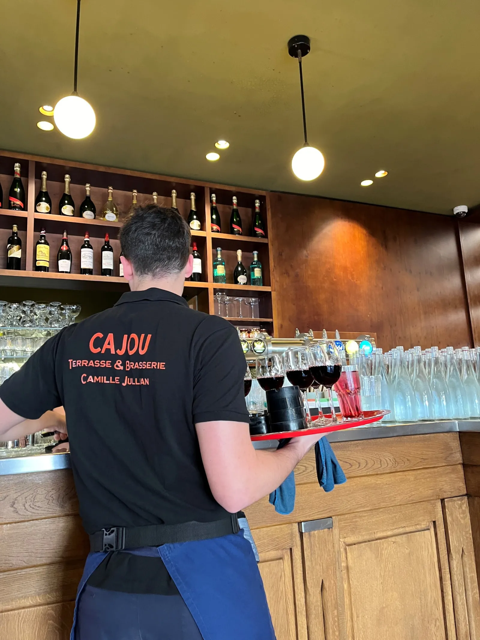 A server wearing a black apron with CAJOU branding works behind the counter of a café terrace and brasserie in Bordeaux, with wine bottle shelves in the background and glasses lined up on the bar.