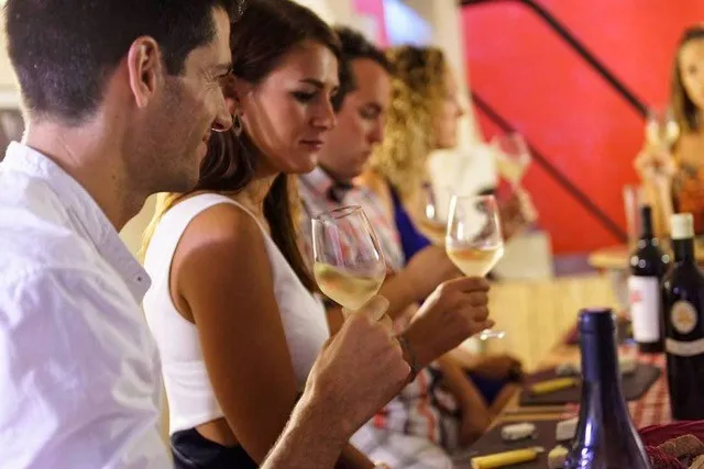 Group of people tasting white wine at a Bordeaux event, holding wine glasses in front of bottles with red décor in the background.