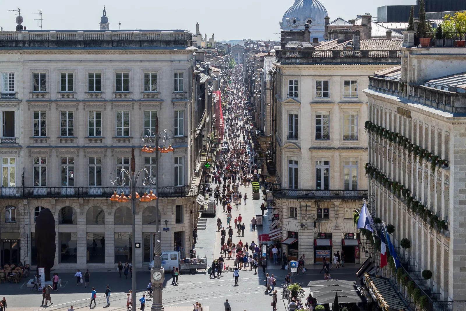 Aerial view of Sainte-Catherine street in Bordeaux, a long pedestrian street lined with Haussmann-style buildings, crowded with pedestrians and a blue-domed basilica in the background.
