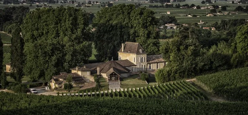 Aerial view of Château Bellefont-Belcier near Bordeaux, surrounded by vineyards and cypress trees, featuring stone buildings with typical French countryside architecture.