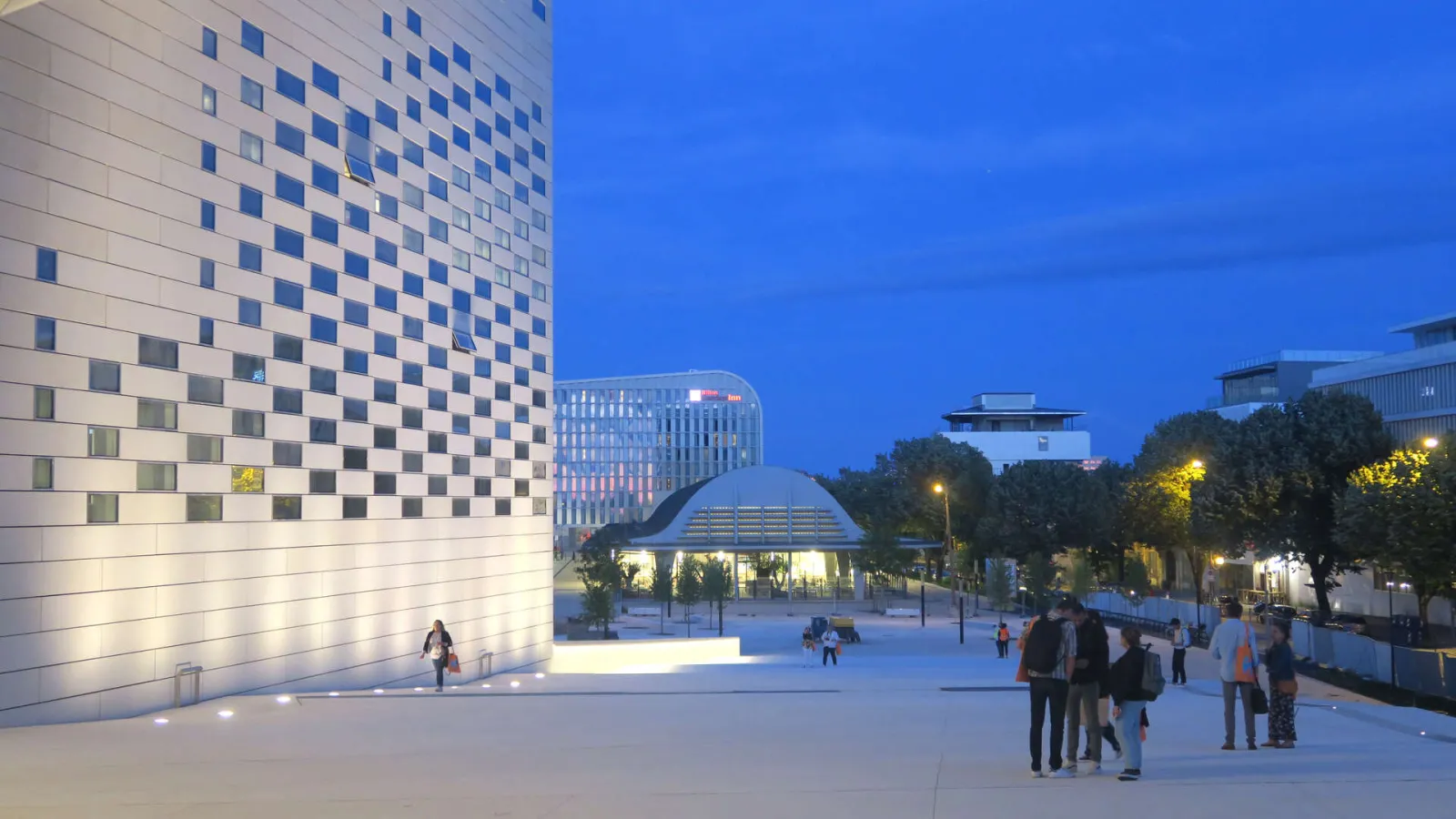 Night view of La Méca in Bordeaux featuring its illuminated white checkered facade, a paved public plaza with visitors, and modern buildings against a twilight sky.