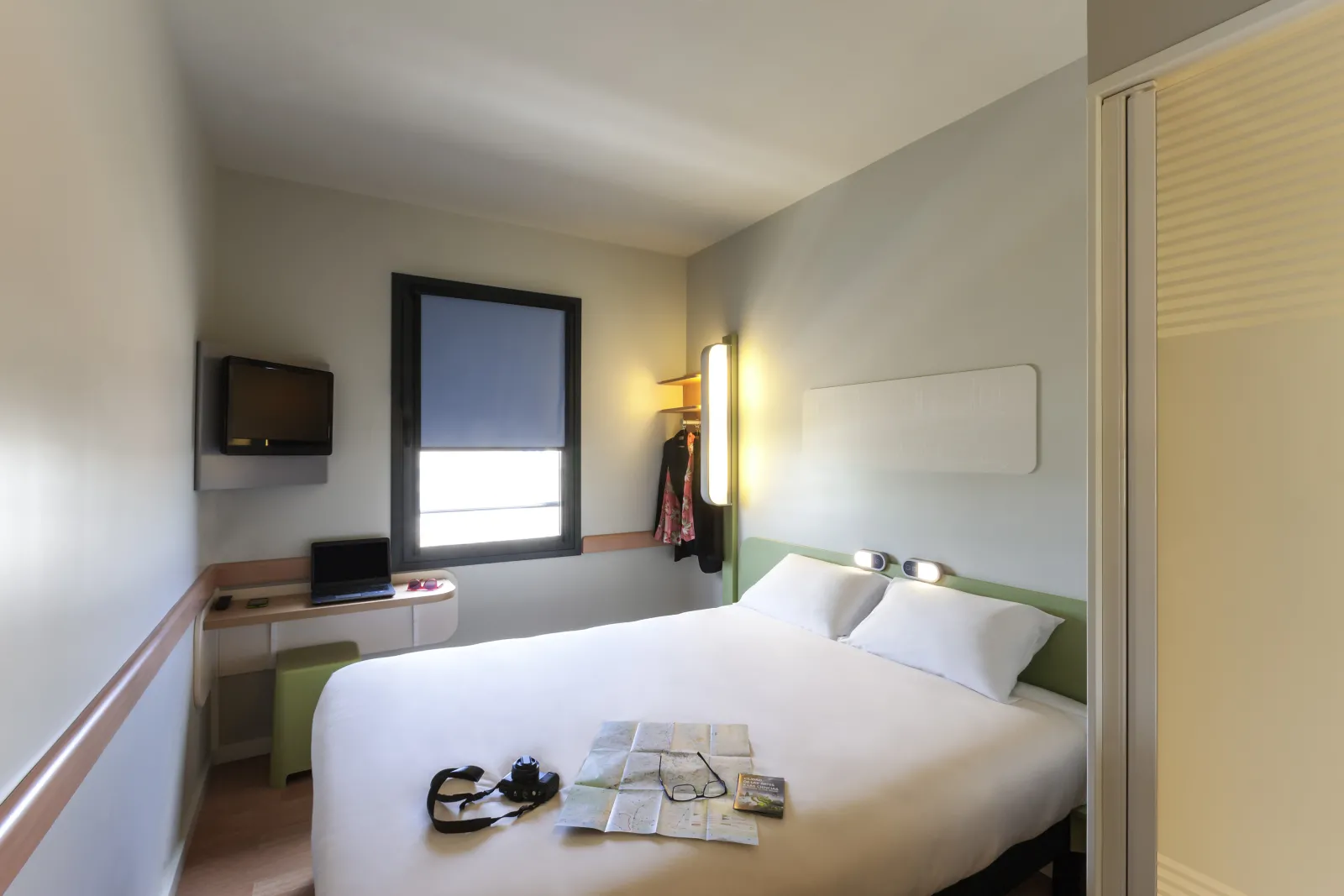 Modern hotel room at Ibis budget Bordeaux Centre Bastide featuring a double bed with white linens, desk with laptop, wall-mounted TV, window with blind, and minimalist storage facilities.