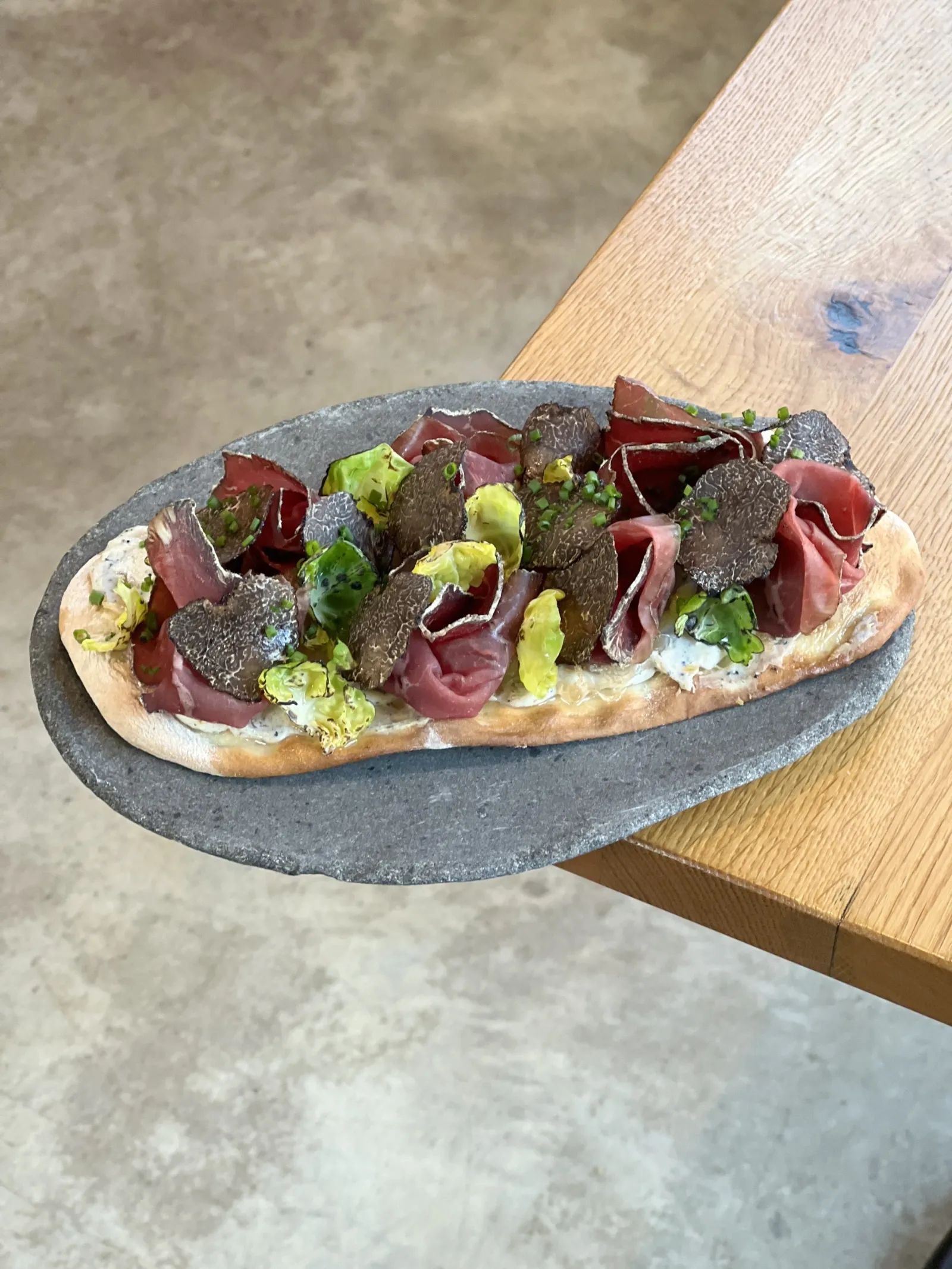 An elongated bread filled with cured meats, green vegetables and white sauce, presented on a grey slate board, on a wooden table in Bordeaux.