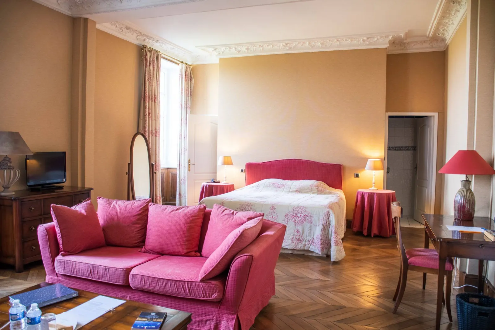 Elegant hotel room featuring a pink sofa, red headboard bed, wooden furniture, ornate white ceiling moldings, and beige walls at Chambres d'Arche in Bordeaux.