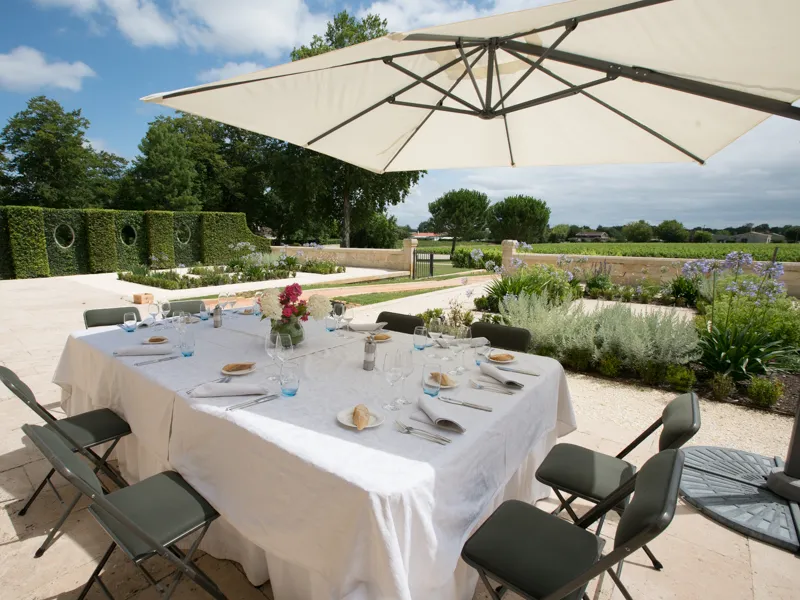 Outdoor dining table set under a white parasol in the gardens of Château Kirwan in Bordeaux, overlooking surrounding vineyards.
