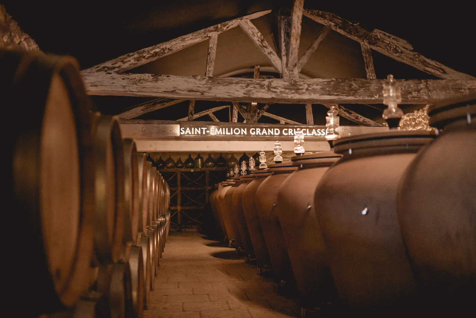 Underground cellar of Château Mangot in Bordeaux featuring rows of oak barrels lined up beneath a massive wooden framework, with Saint-Émilion Grand Cru signage visible