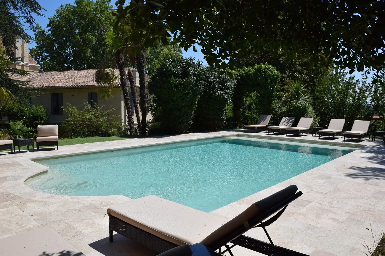 Rectangular outdoor swimming pool with turquoise water surrounded by loungers and sunbeds, facing a Provençal-style building surrounded by lush vegetation at Château Bellefont Belcier in Bordeaux.
