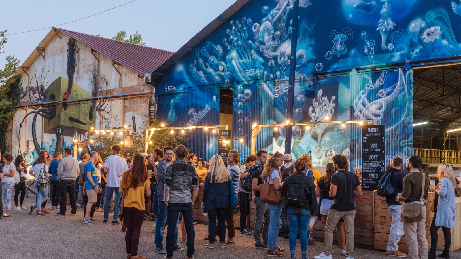 A crowd of visitors gathers in the evening in an outdoor courtyard decorated with street art and string lights, featuring buildings with blue and white marine-themed murals, at Darwin Caserne Niel in Bordeaux.