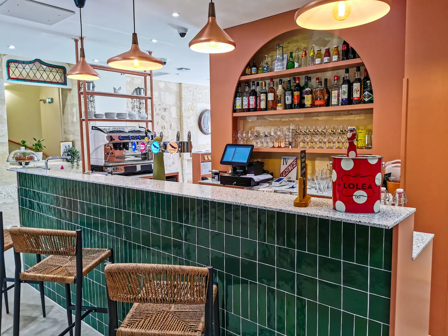 Modern bar counter with green tile cladding, shelves of alcohol bottles, espresso machines, wicker stools and copper pendant lights in a Bordeaux establishment.