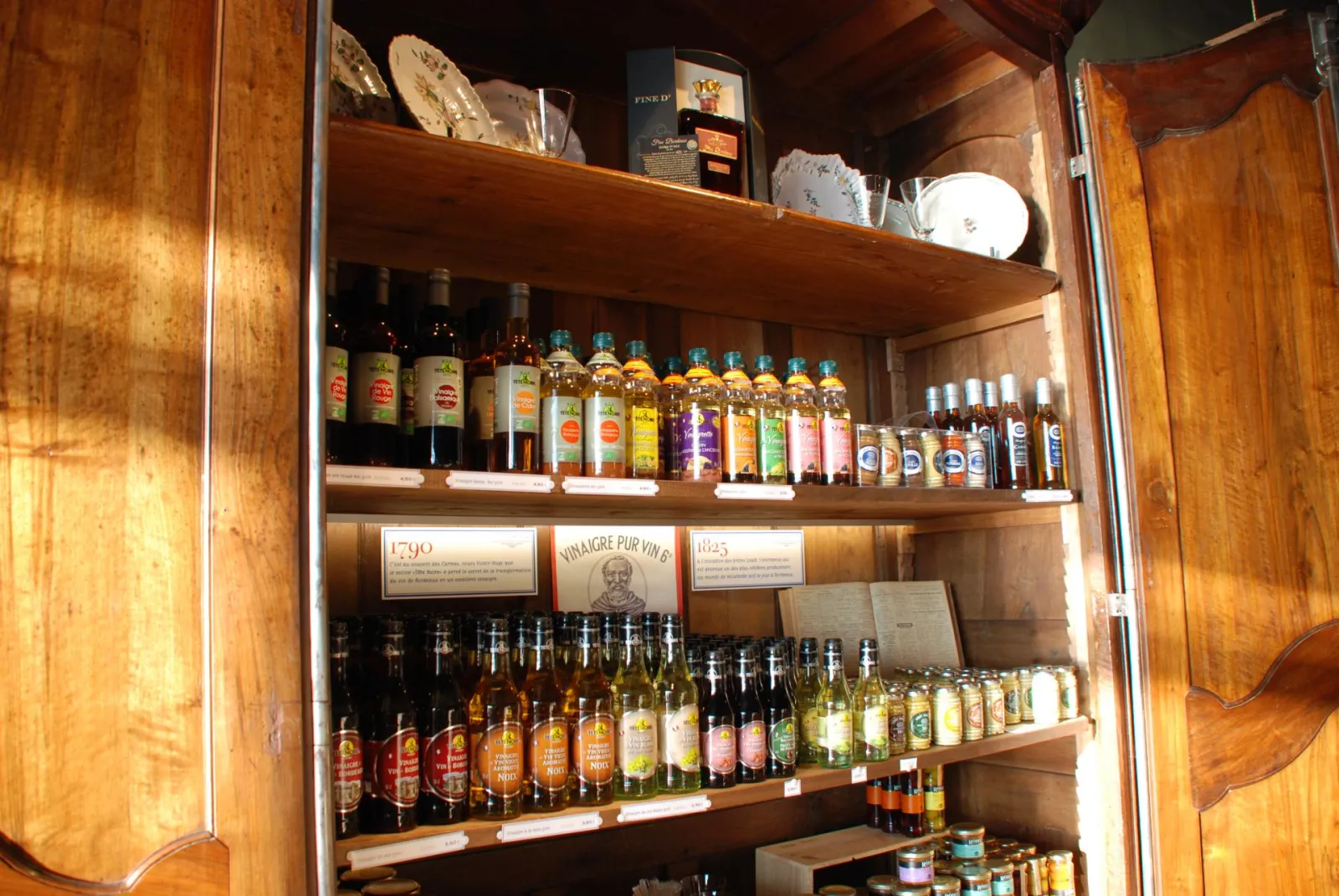 Open wooden cabinet displaying a collection of vinegar and spirit bottles arranged on three shelves, with historical plaques dated 1790, 1825 and French inscriptions, in a Bordeaux shop.