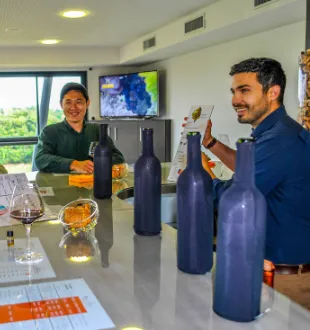 Two men taste wine in a cellar at Château La Croizille in Bordeaux, with blue wine bottles and filled glasses on the counter.