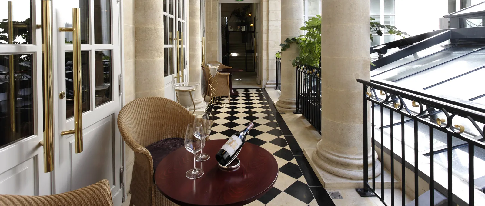 Elegant hallway of Le Pressoir d'Argent Gordon Ramsay restaurant in Bordeaux, featuring black and white checkered flooring, stone columns, gilded and white doors, beige armchairs, and a burgundy round table with wine glasses and bottle