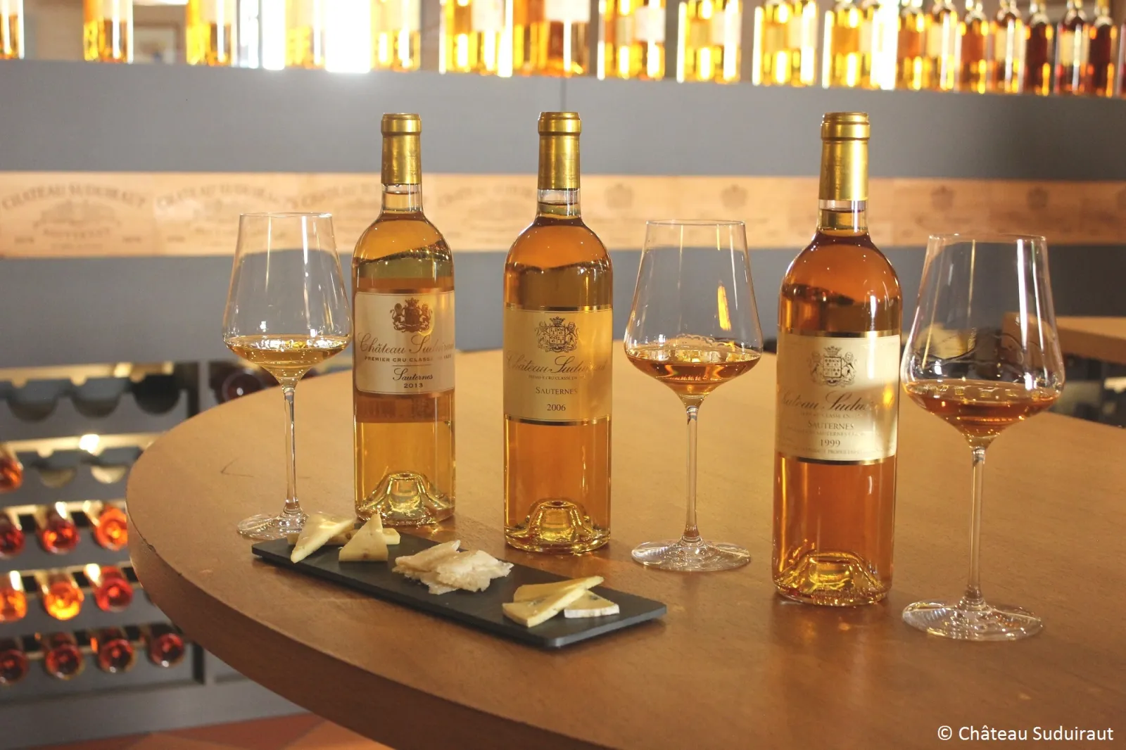 Tasting of Château Suduiraut wines: three bottles of Sauternes with filled glasses and a cheese tasting board, in a cellar with illuminated bottles.