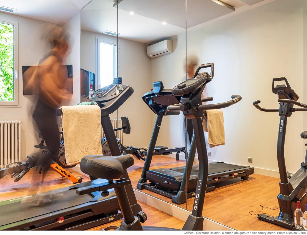 Modern fitness room at Château Bellefont Belcier in Bordeaux featuring multiple cardio equipment including treadmills, stationary bikes and elliptical machines, wooden flooring, wall mirrors and yellow towels