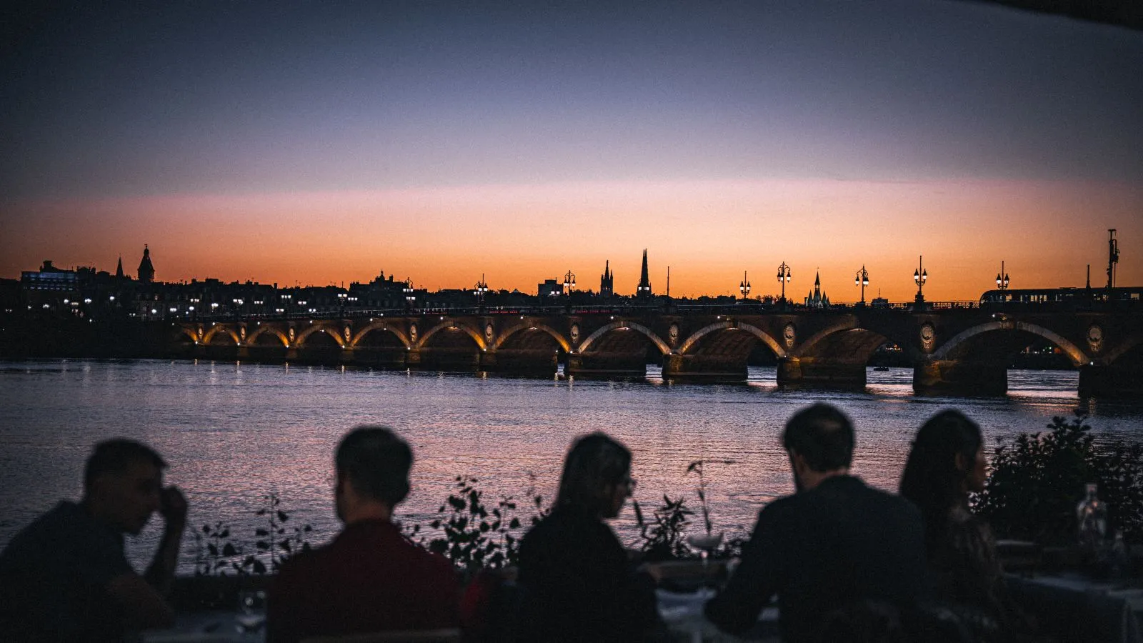 Twilight view of Bordeaux's Stone Bridge with illuminated arches, historic skyline in the background, and people in the foreground watching the sunset over the Garonne River.