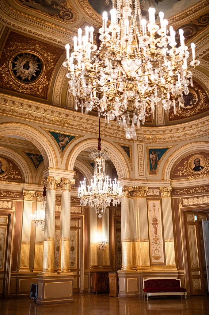 Opulent baroque room featuring ornate gilded ceiling with coffers, Corinthian columns, wall frescoes, and two illuminated crystal chandeliers.