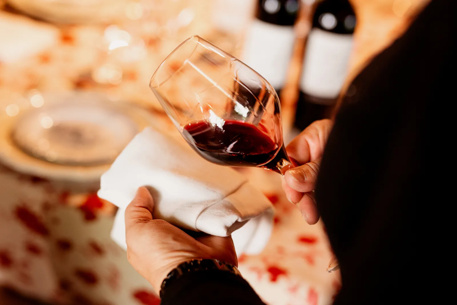 A hand holding a glass of Bordeaux red wine during a tasting, with bottles and glasses in the background