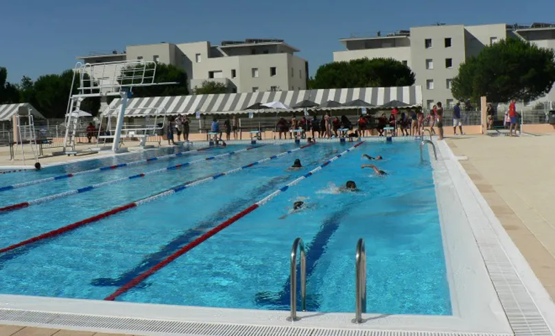 Floirac municipal swimming pool in Bordeaux with multiple swimmers in lanes marked by red and blue lane dividers, surrounded by spectators and residential buildings in the background.