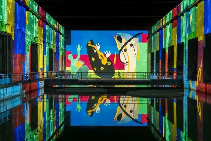 Immersive digital installation at Bordeaux's Bassins des Lumières featuring vibrant abstract figures inspired by Matisse, with bright colors (blue, red, yellow, green) projected on walls and reflected in water.