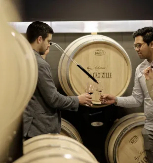 Two men examining a wine barrel in the cellar of Château La Croizille in Bordeaux, surrounded by wooden casks.