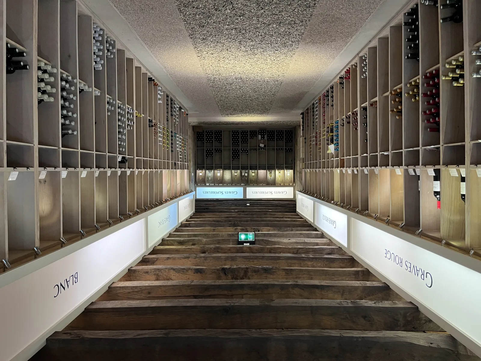 Interior of the Maison des Vins de Graves in Bordeaux, display gallery featuring wine bottles arranged in wooden racks on both sides of a corridor, with textured ceiling and ambient lighting.