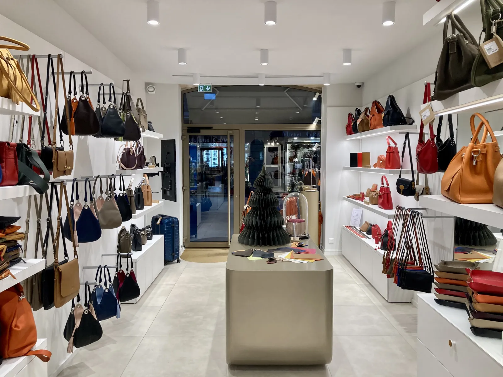 Interior of a leather goods shop in Bordeaux displaying colorful handbags on white shelves and wall displays, with a black decorative tree in the center and glass entrance visible.