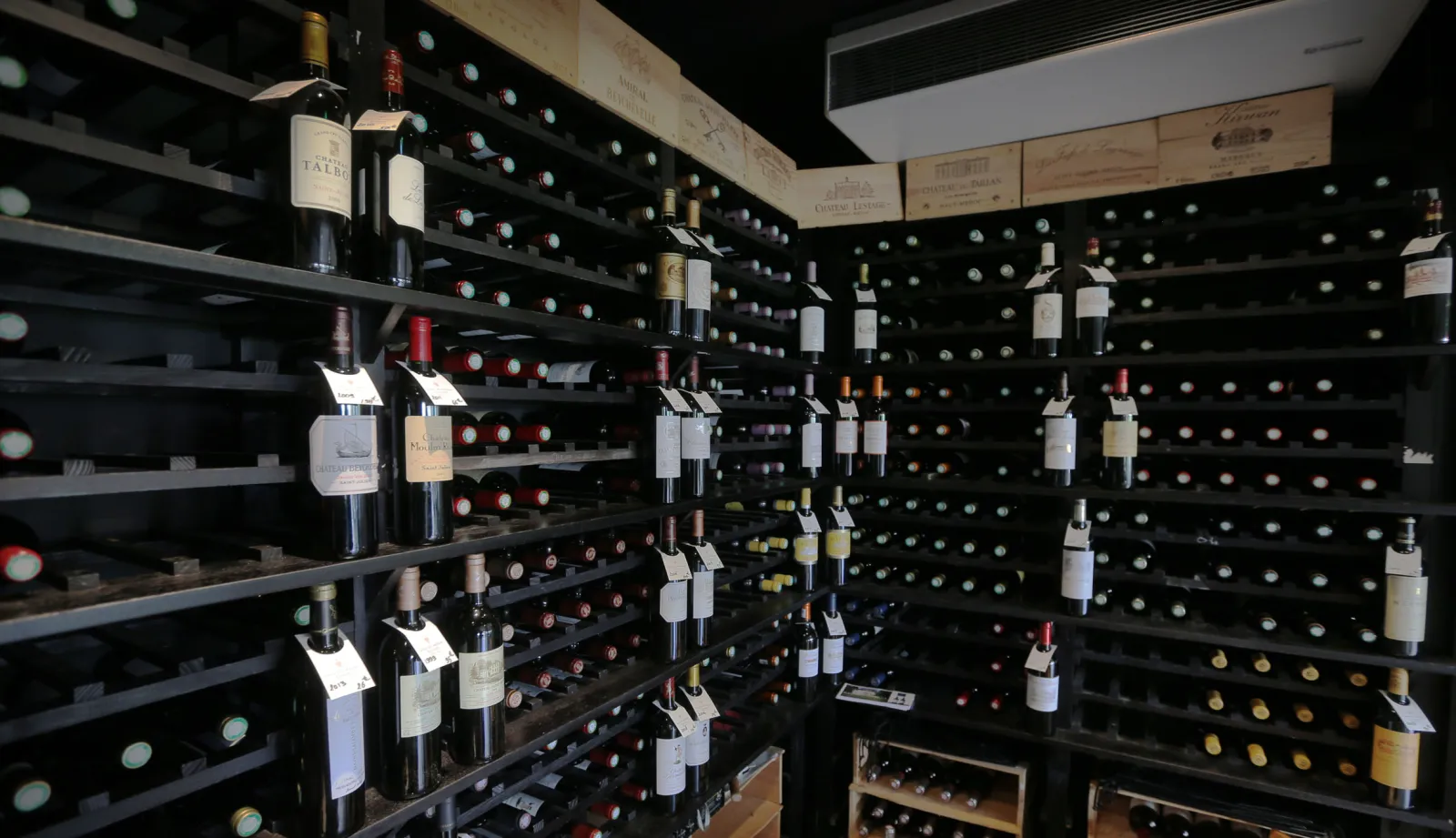 Wine cellar with black shelving displaying hundreds of wine bottles organized with white labels, and wooden wine crates visible on the ceiling, at Le Club restaurant in Golf du Pian Médoc near Bordeaux.