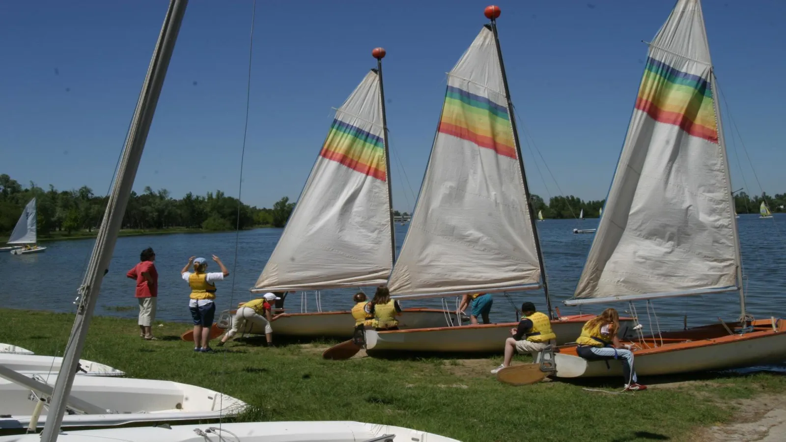 Three sailboats with white sails and rainbow stripes beached on grass with children in yellow life jackets preparing the sails at Lac Beach in Bordeaux