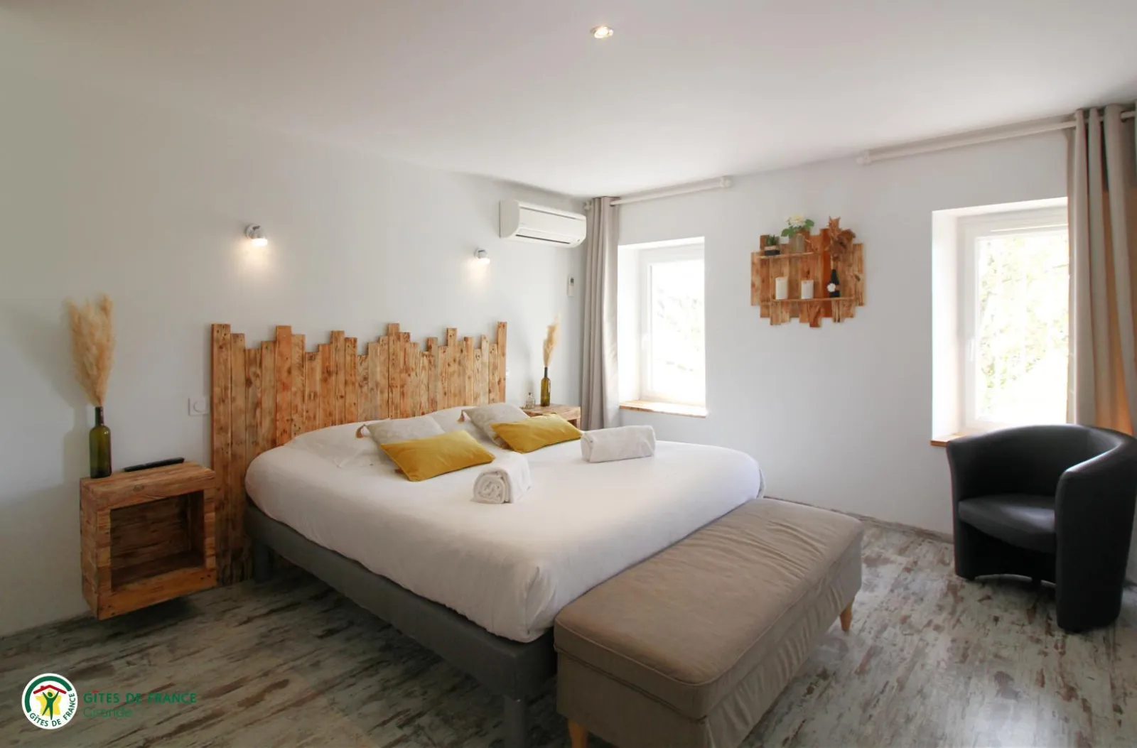Bright modern bedroom featuring a double bed with white linens, wooden headboard, yellow pillows, windows overlooking the garden, black armchair, wooden nightstand and minimalist decor.