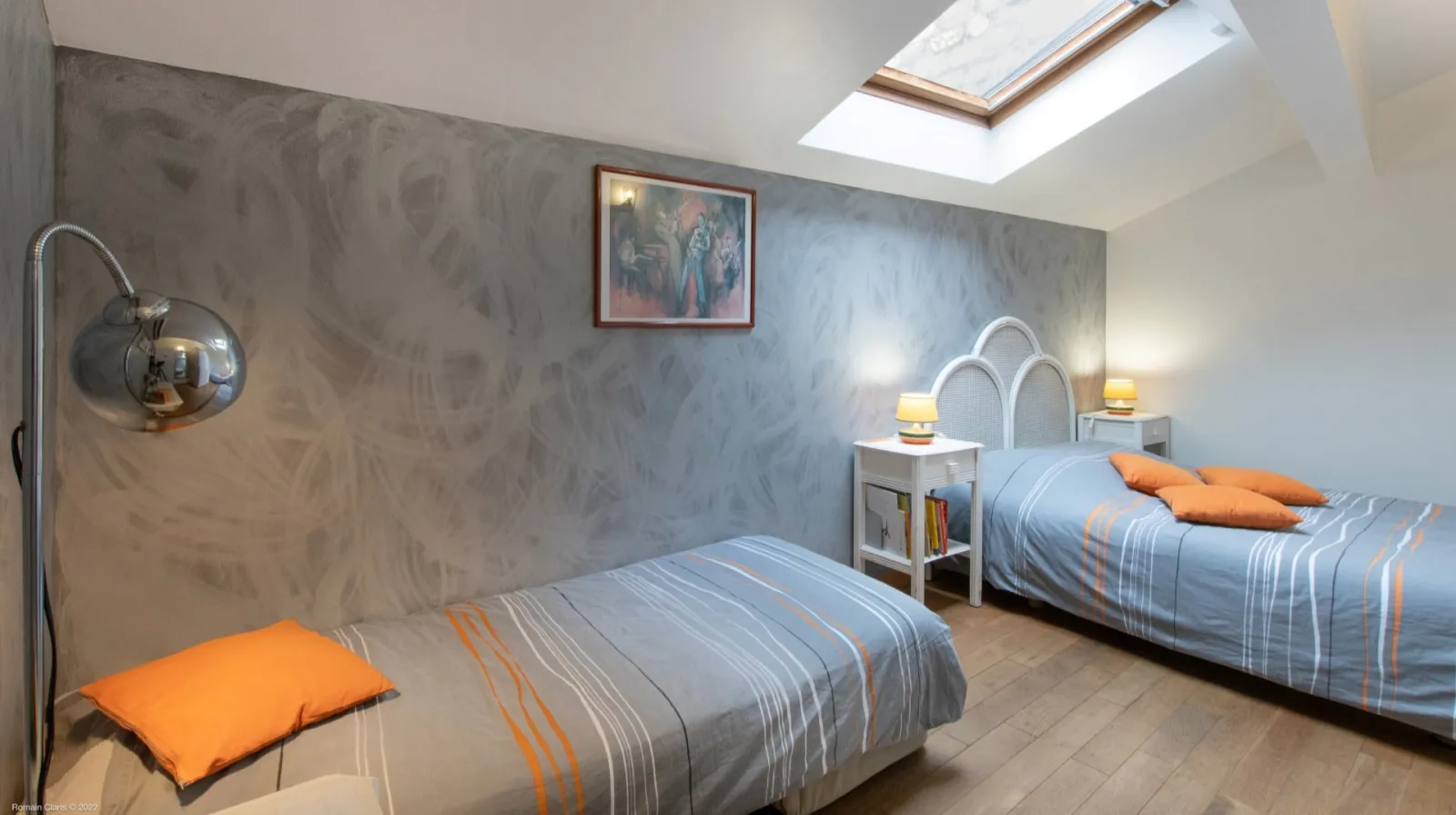 Guest bedroom in Bordeaux featuring two twin beds with blue and orange striped linens, chrome arc lamp, light gray walls, skylight, white bedside tables, and minimalist decor.