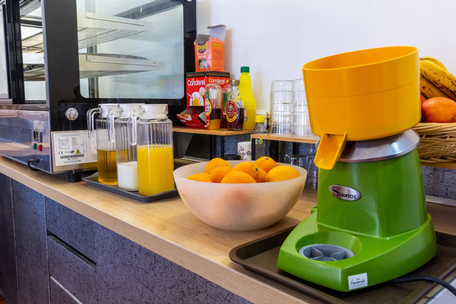 Breakfast counter featuring a green juice maker, bowl of oranges, orange juice containers, bananas, refrigerator and condiment bottles at Hôtel Notre Dame in Bordeaux.