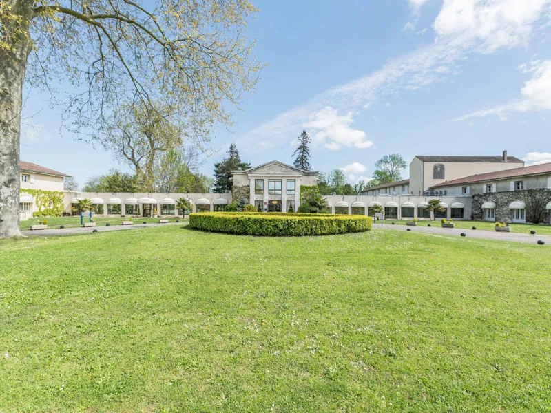 Front view of Hôtel Le Relais de Margaux featuring stone buildings, expansive green lawn, manicured hedges, mature trees, and blue cloudy sky.