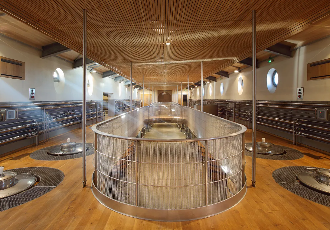 Modern winery interior featuring a large stainless steel circular vat in the center, surrounded by wooden oak barrels, wooden slatted ceiling and natural light at Château Soutard in Bordeaux.
