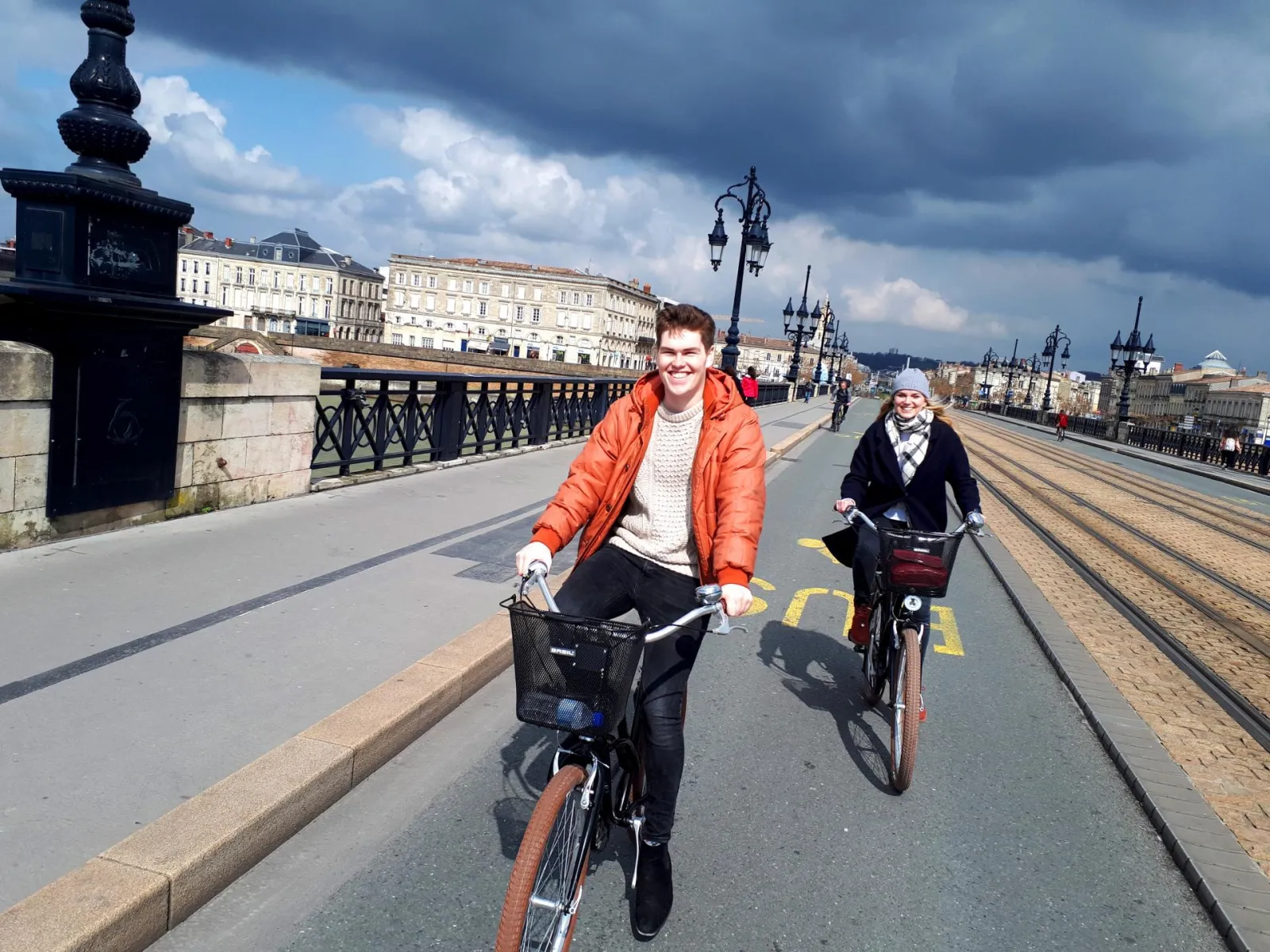 Two smiling cyclists riding bikes on a dedicated bike lane crossing a historic bridge in Bordeaux, with classical buildings and a cathedral in the background