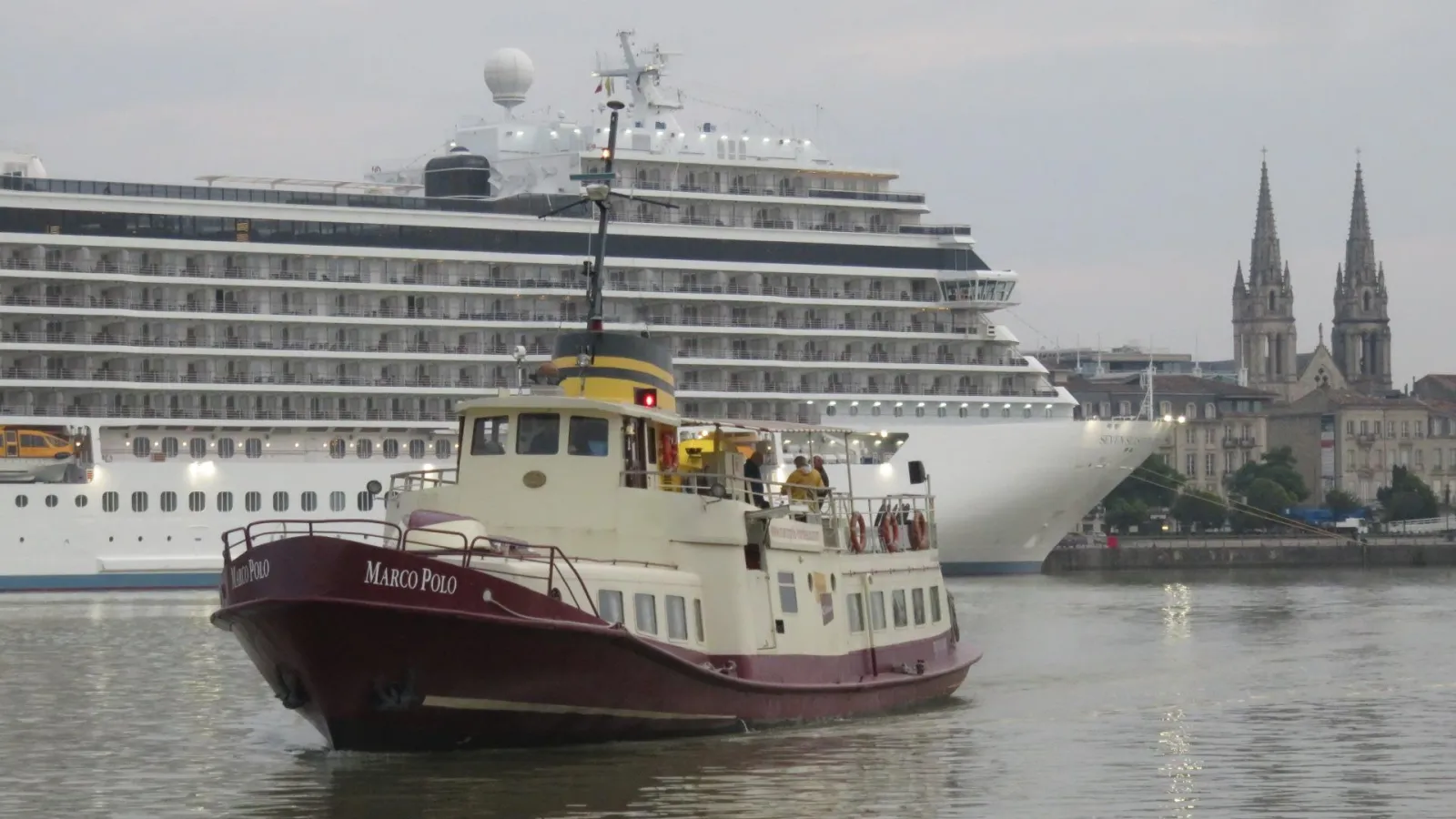 The river cruise ship Marco Polo sails on the Garonne in Bordeaux, with a large cruise ship in the background and Saint-André Cathedral visible on the riverbank.