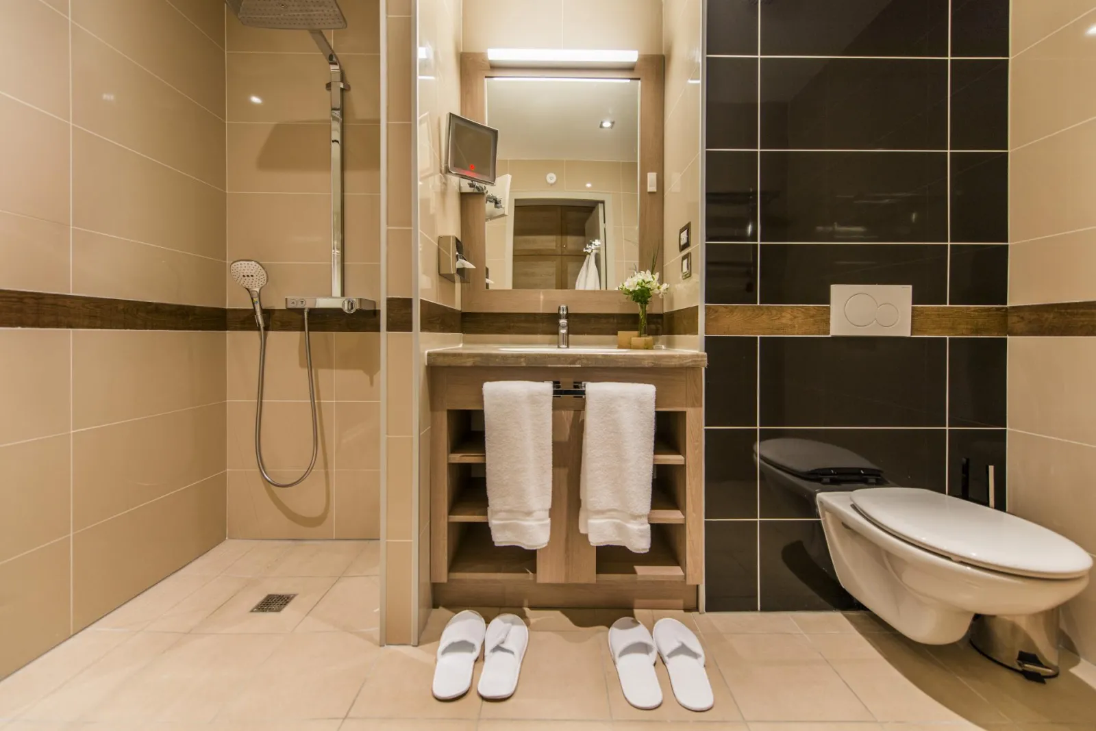 Modern bathroom in Hotel Vatel Bordeaux featuring walk-in shower, double sink vanity, suspended toilet, beige and black tiles, illuminated mirror, white towels and white slippers