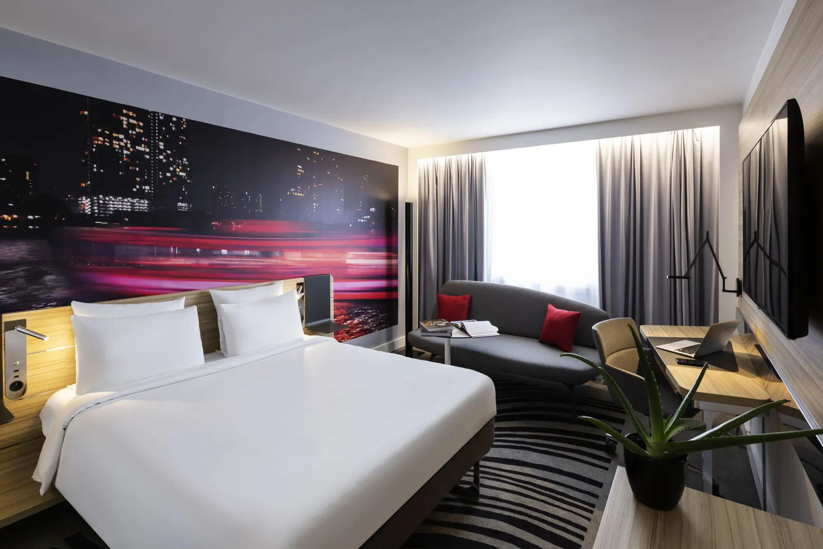 Modern hotel room at Novotel Bordeaux Centre featuring a king-size bed with white linens, a night cityscape wall mural in red and gold tones, grey sofa with red cushions, floor-to-ceiling grey curtains, and contemporary amenities.