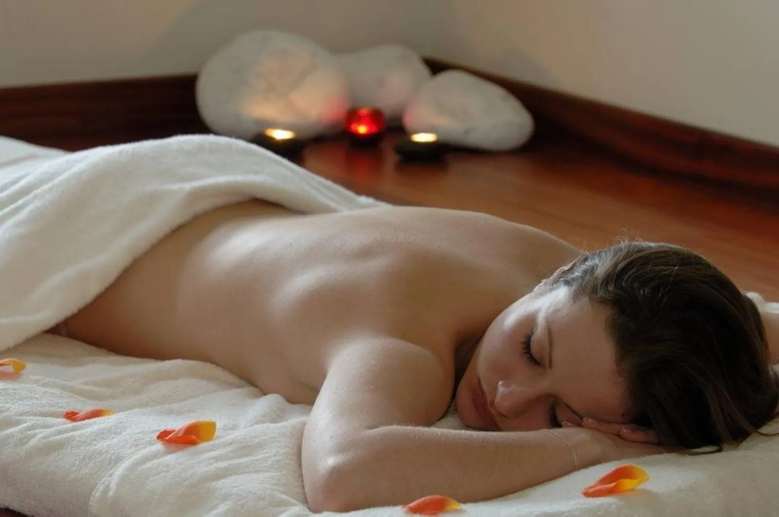 Woman lying face down on a white bed with orange rose petals, in a spa atmosphere with lit candles and heated stones in the background