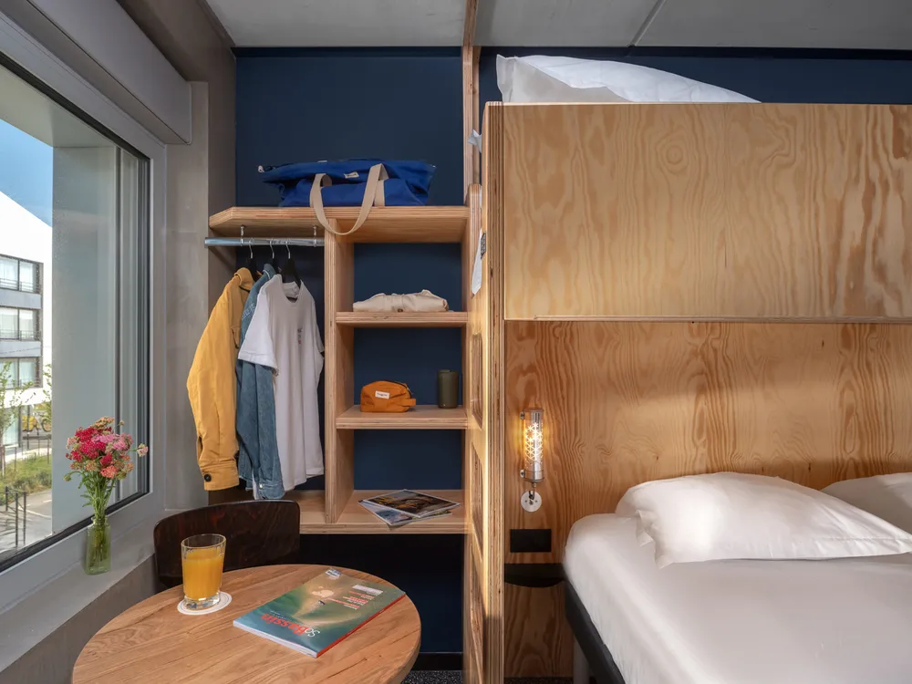 Compact and modern hotel room featuring a single bed, round desk by the window, integrated wooden storage with shelves and wardrobe, and minimalist décor in navy blue and light wood tones.